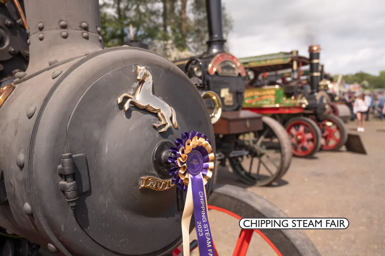 Close-up of a decorated steam engine boiler with a brass horse emblem and purple award ribbon, with vintage steam engines and crowds visible behind.