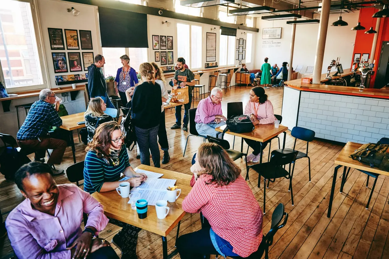 Adults gather at tables in a spacious community hall with wooden floors, drinking beverages and chatting during a social event.