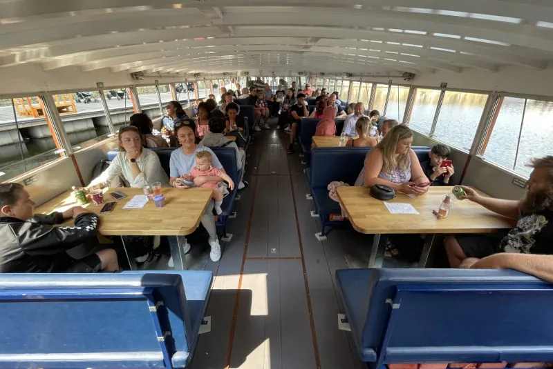 Interior of a passenger ferry or tour boat with adults and children seated at tables along both sides, dining and enjoying the voyage on water.