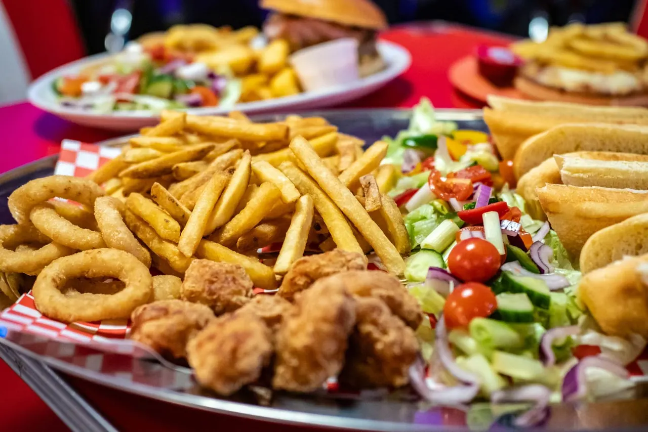 A table laden with fast food including fried chicken, chips, onion rings, hot dogs and salads on red and white checked paper.