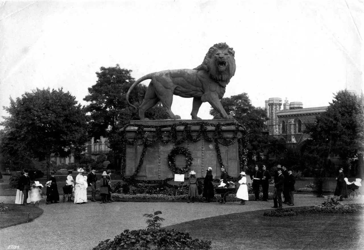 Large stone lion statue on a pedestal surrounded by visitors in early twentieth-century dress, with buildings and trees in background.
