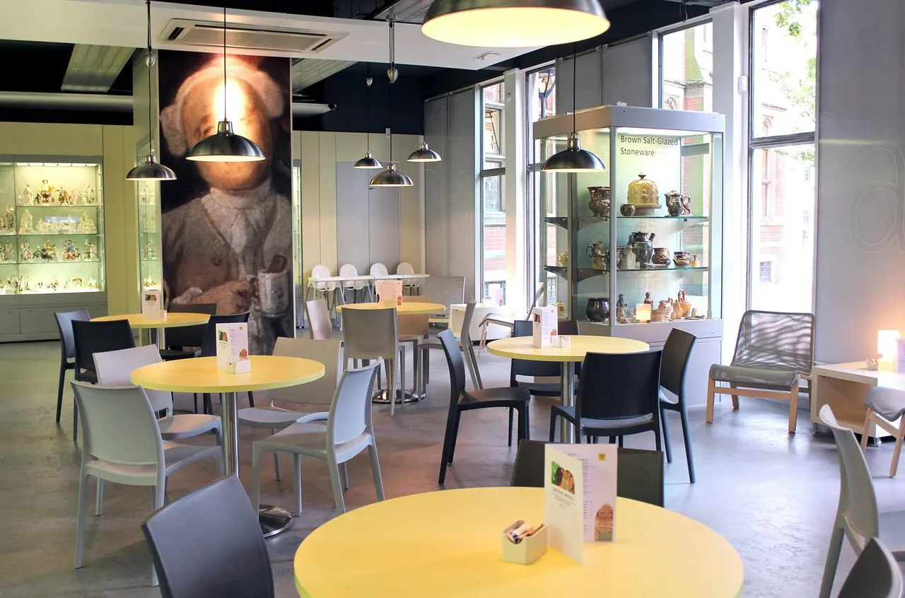 Modern café interior with yellow round tables, black and white chairs, pendant lights, display cabinets of pottery, and large windows overlooking the street.