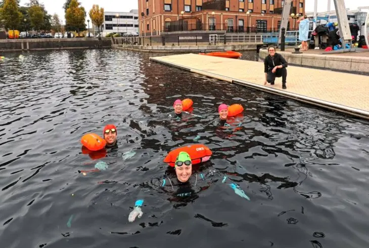 Swimmers in orange buoyancy aids and goggles participate in a supervised open-water swimming session in a dock basin.