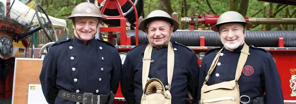 Three men in vintage firefighter uniforms and helmets stand in front of red fire engines and equipment.