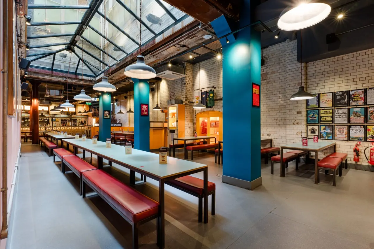 Industrial-style dining hall with long communal tables, red bench seating, blue support columns, pendant lighting and exposed brick walls.