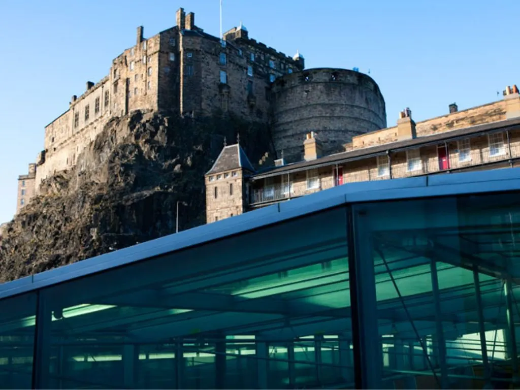 Historic castle on rocky outcrop with modern glass-fronted building in foreground under clear blue sky.