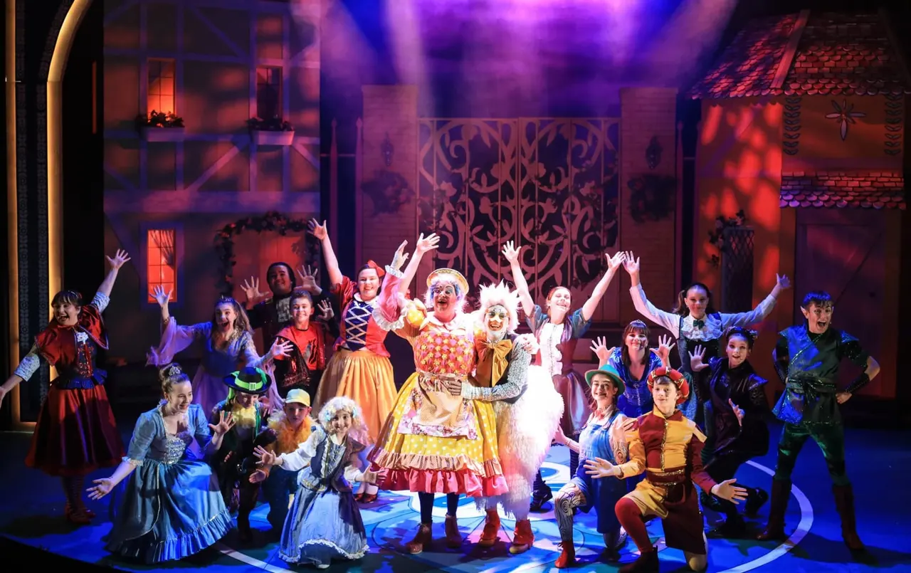 Cast of pantomime performers in elaborate colourful costumes on stage, arms raised in celebration under purple and red stage lighting.