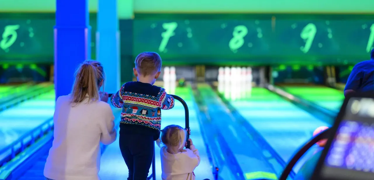 Three young children standing between bowling lanes with brightly lit pins and numbered lane markers visible.