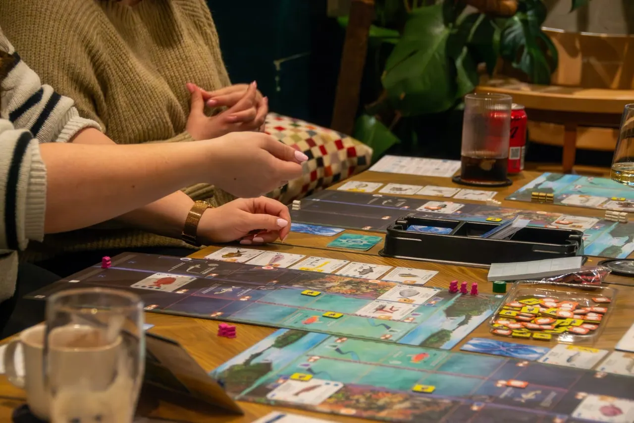 Person playing a board game at a wooden table with drinks and game pieces.