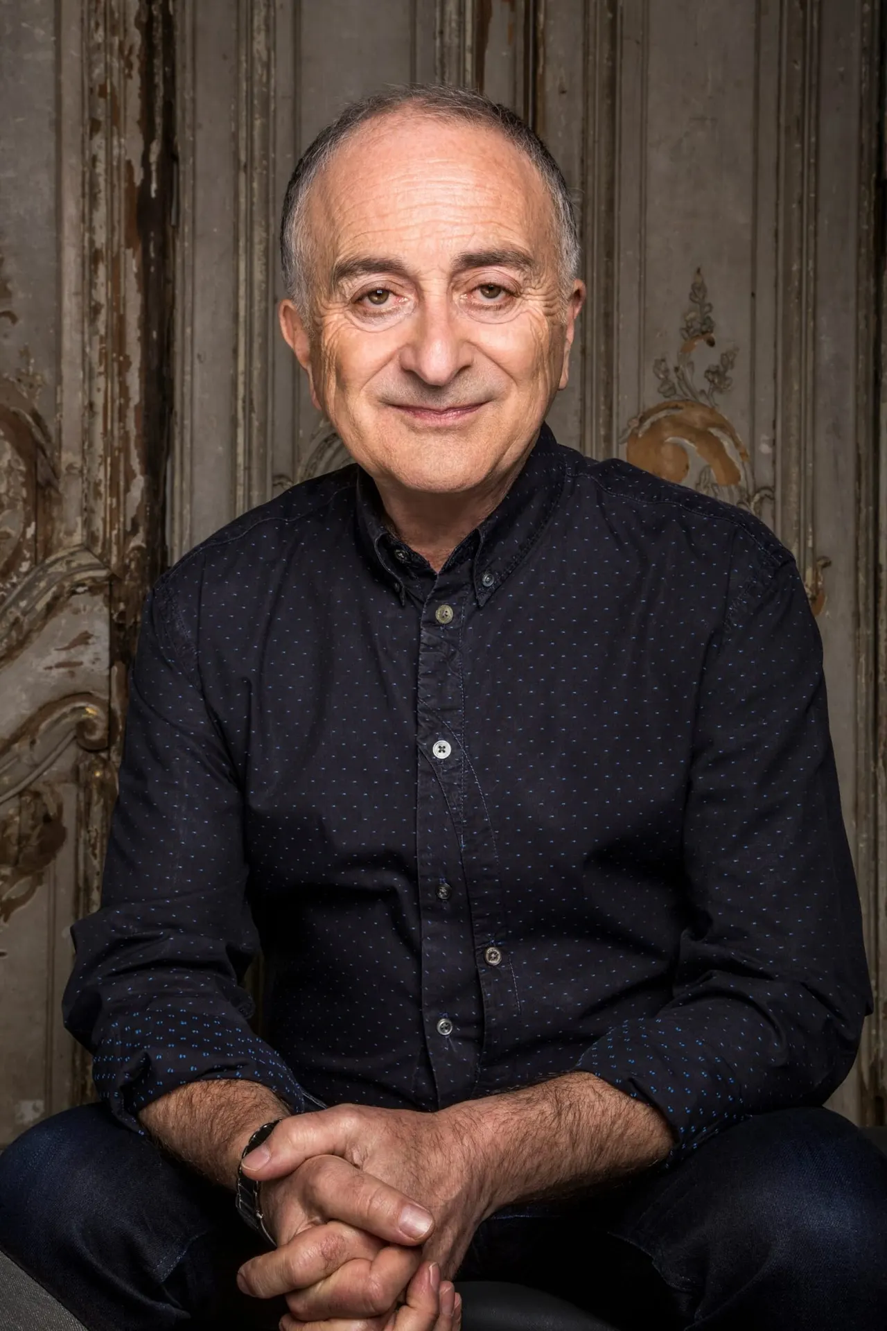 This is a portrait photograph of a middle-aged man sitting in front of a weathered, ornate backdrop, wearing a dark navy button-up shirt.