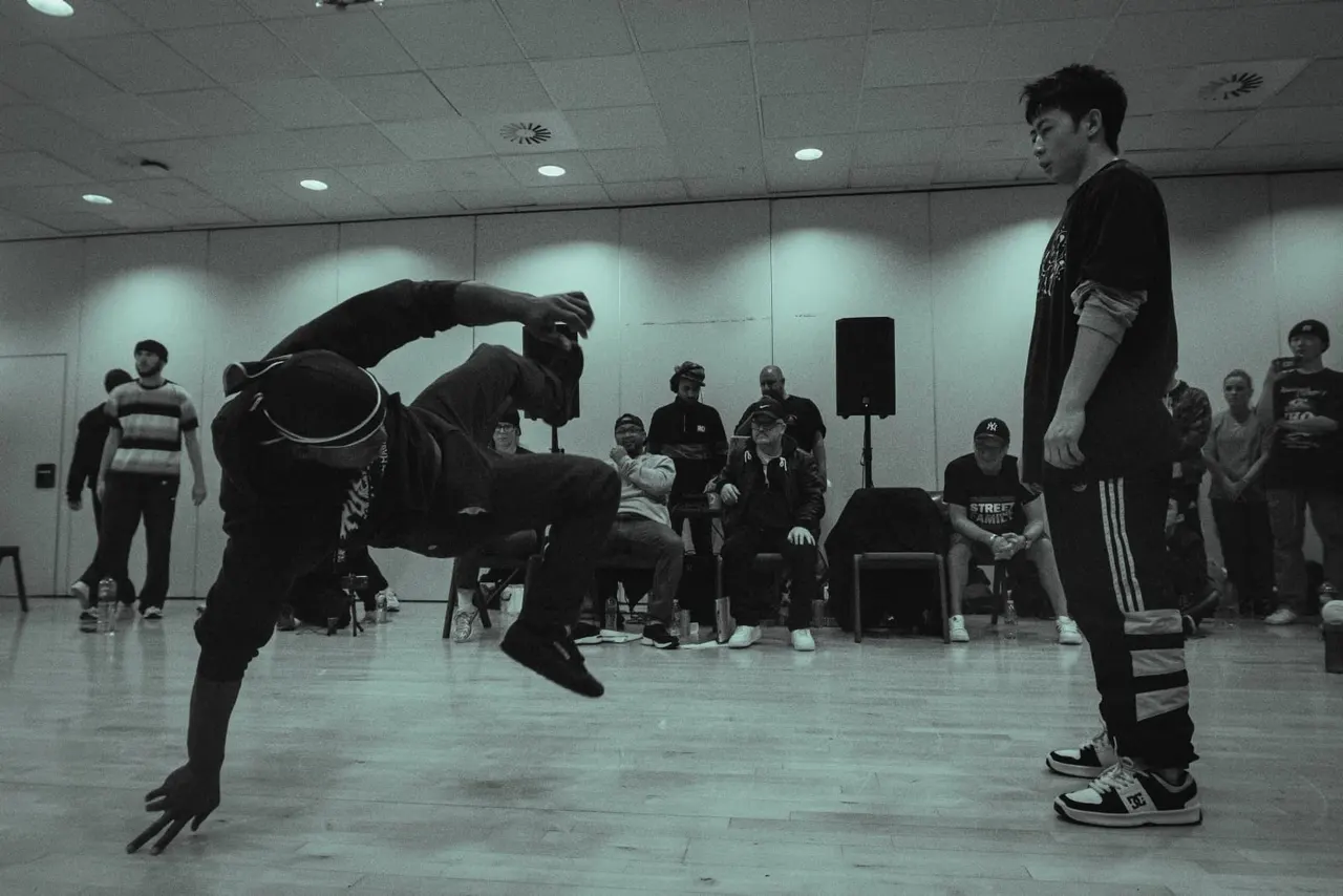 Breakdancer performing a backflip in an indoor studio whilst spectators watch from chairs around the dance floor.