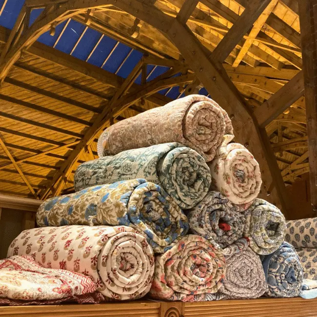 Rolled carpets and rugs stacked inside a wooden barn structure with exposed beams and skylight openings.