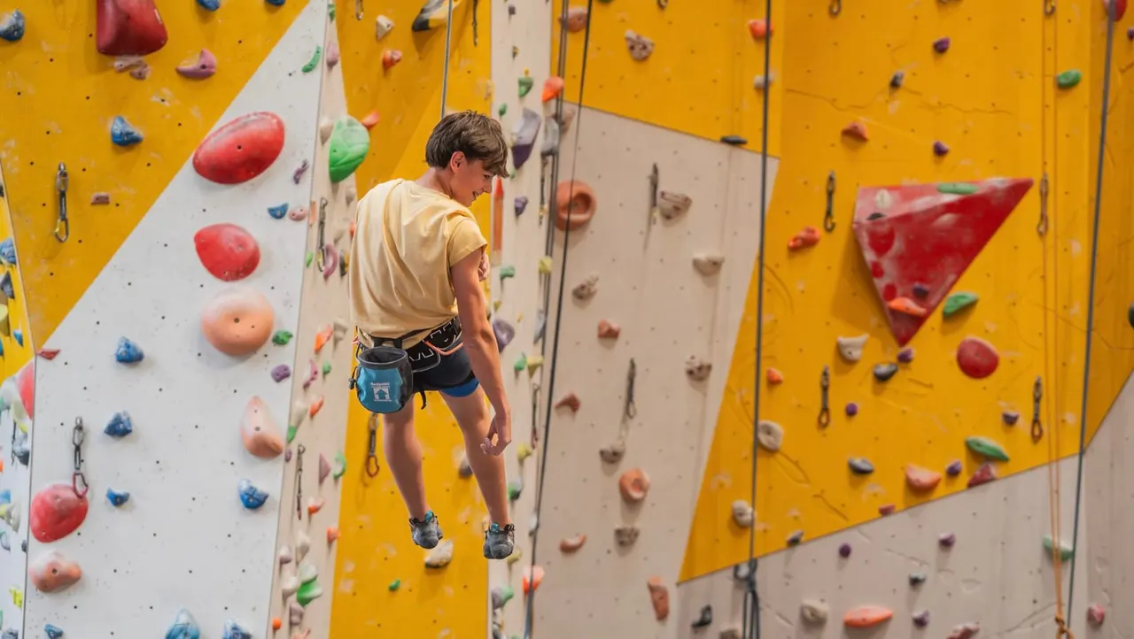 Child rock climbing on an indoor wall with coloured handholds whilst secured by a rope.
