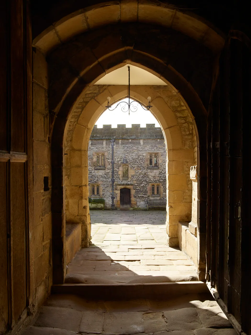 View through a stone archway into a castle courtyard, showing a historic stone building with crenellated walls and mullioned windows beyond.