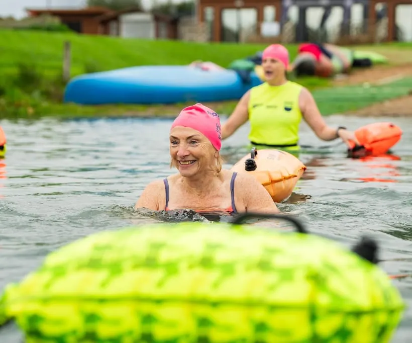 Adult swimmers wearing buoyancy aids and swim caps in an outdoor lake with inflatables and instructors nearby.