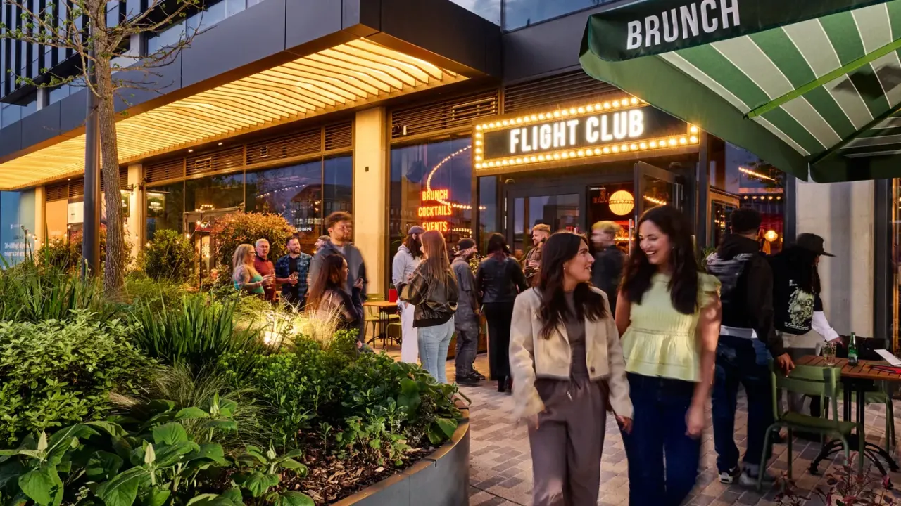 Groups of people socialising outside a brightly lit Flight Club venue at dusk, with planted landscaping and a green brunch canopy visible.
