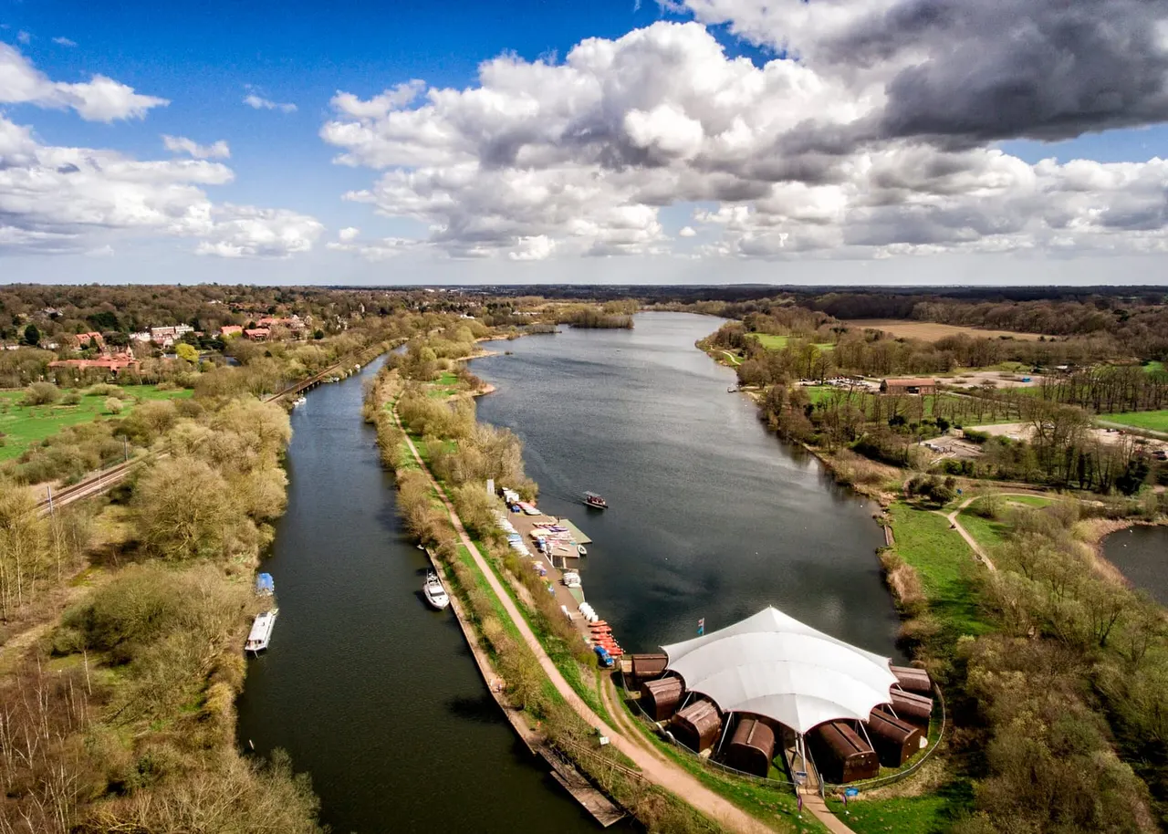 Aerial view of a riverside venue with a white marquee, moored boats, and landscaped grounds beside a wide river valley.
