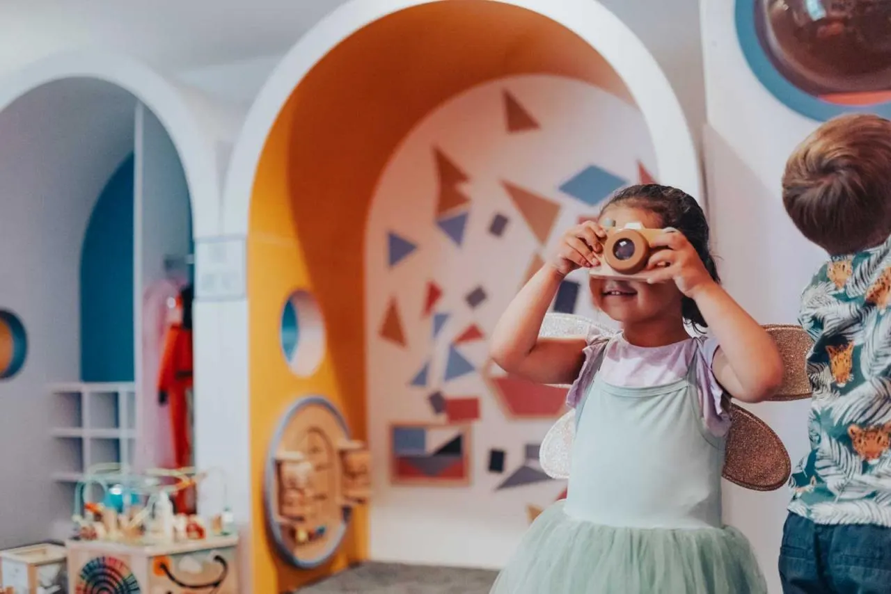 Child holding a toy camera up to face inside a colourful, geometric-patterned play space with another child nearby.