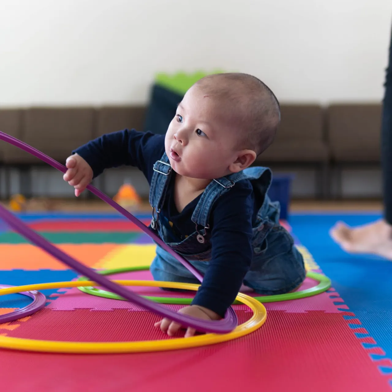 Toddler crawling over coloured hoops on a padded play mat during an activity session.