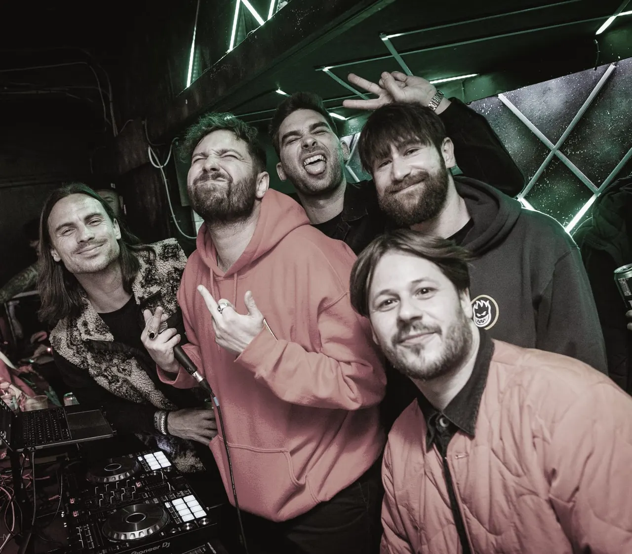 Five men posing together at a nightclub with DJ equipment and neon green lighting visible behind them.