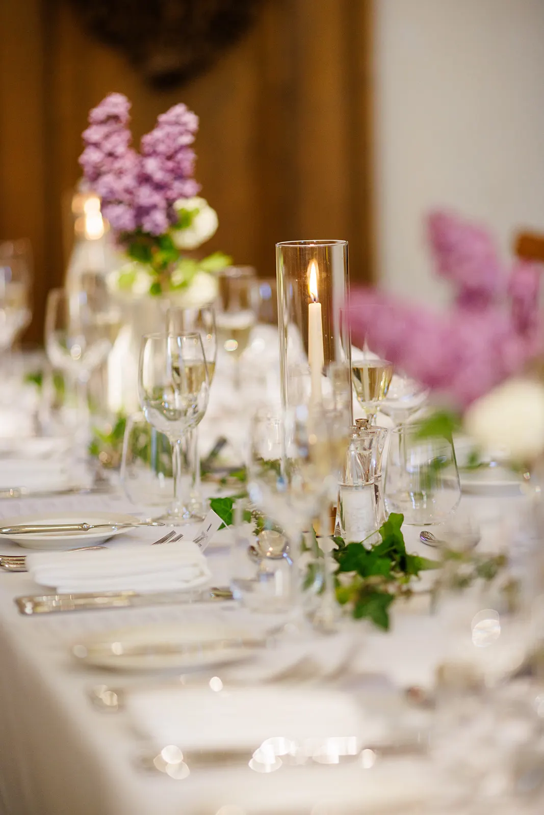 Formal dining table set with glassware, cutlery, white napkins, lit candles in glass holders and pink and white floral centrepieces.