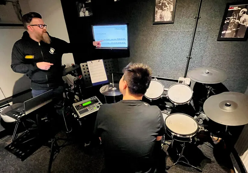 Instructor teaching a student at an electronic drum kit in a music lesson studio.