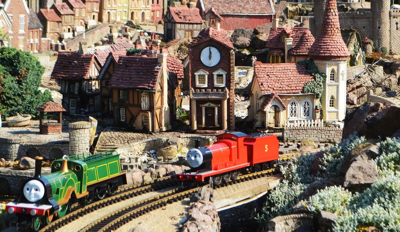 Miniature model railway with green and red trains running through a detailed brick village with clock tower and period buildings.