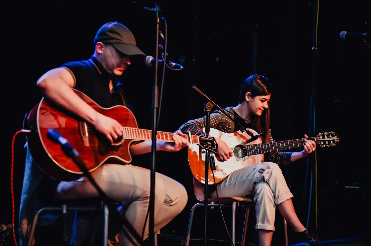 Two musicians performing with acoustic guitars on stage under blue lighting.