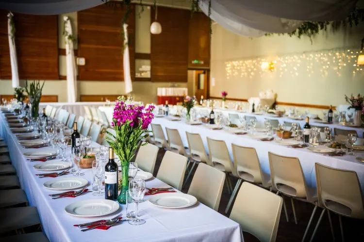 Long banquet tables with white tablecloths, place settings, and pink flower arrangements in an elegant event space with warm lighting.