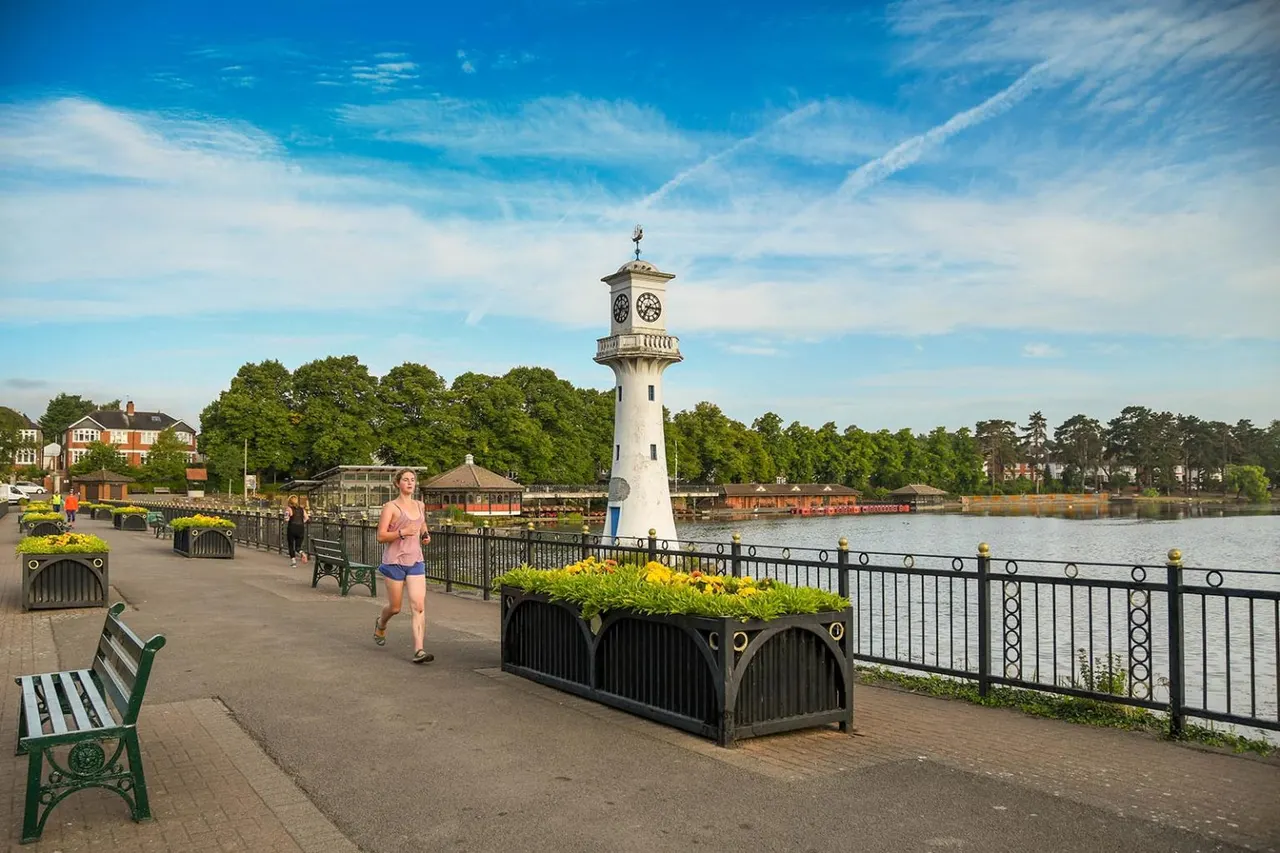 A jogger runs along a waterside promenade past a white clock tower and Victorian boating lake, with benches and flower planters lining the path.