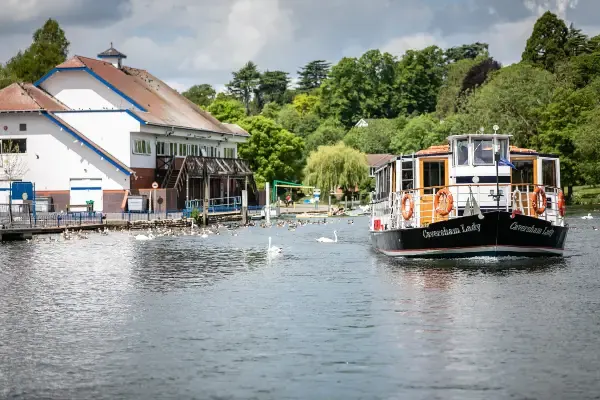 A riverboat with orange and white livery on the Thames with a riverside building and swans nearby.