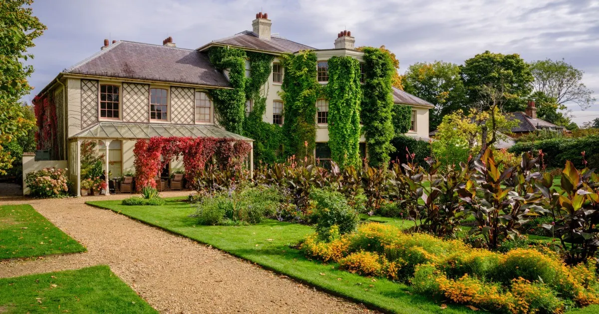Historic country house with ivy-covered facade and manicured gardens featuring gravel pathways and autumn-flowering borders.