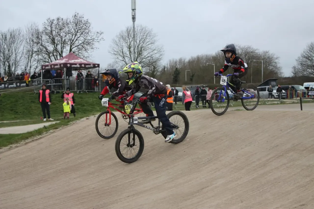 Young BMX riders competing on a dirt track with spectators and gazebo in the background.