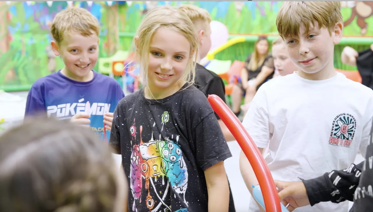 Three children smiling at an indoor soft play centre with colourful play equipment visible in the background.