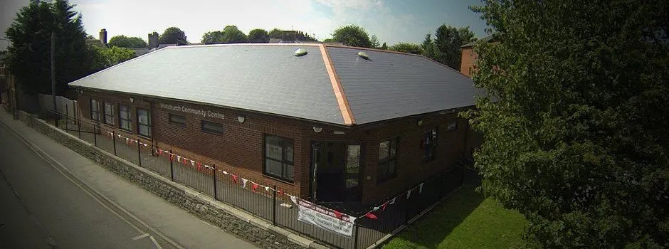 Brick community centre building with light-coloured roof, viewed from street with surrounding greenery and fencing.