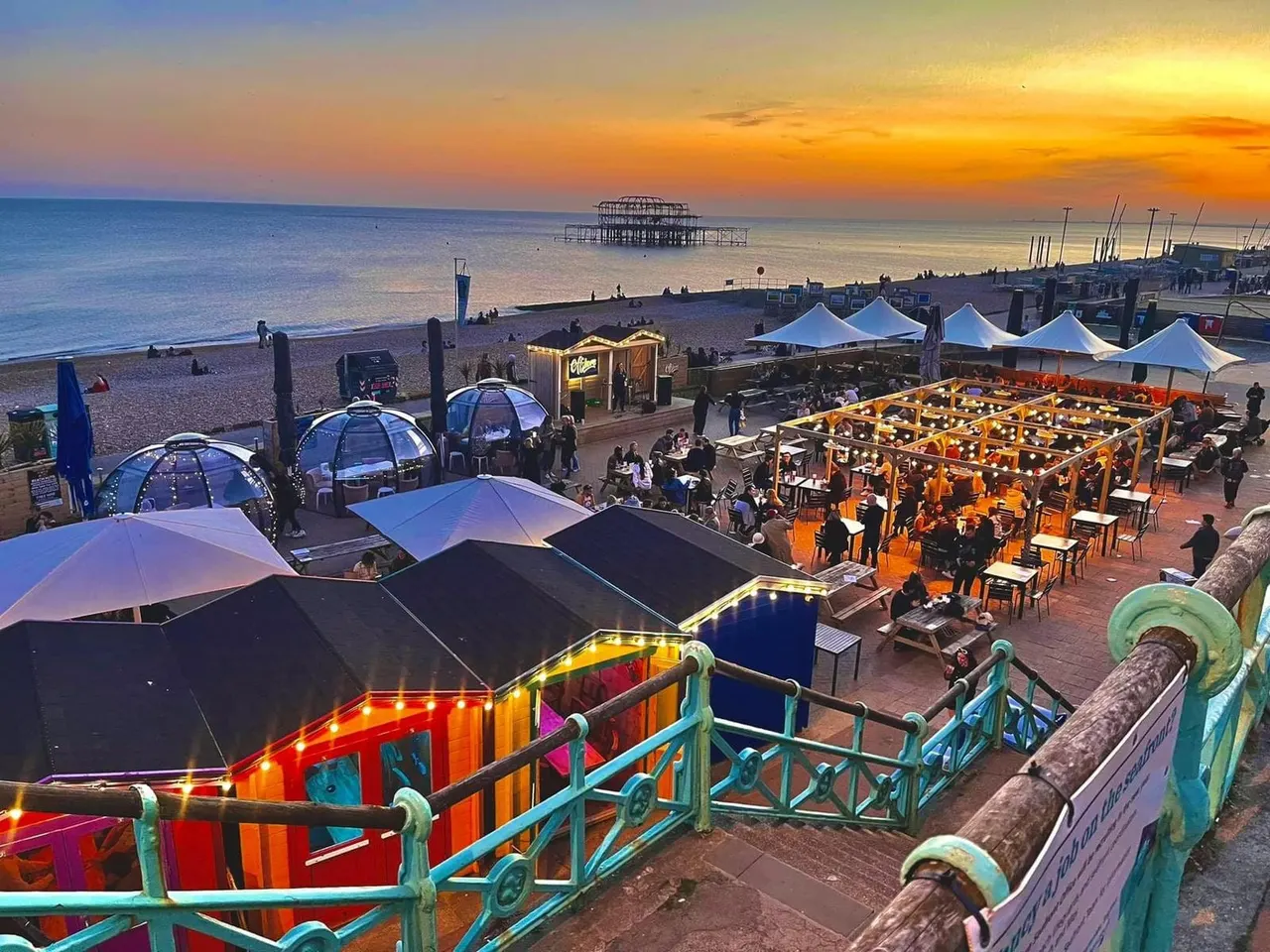 Sunset view of a beachfront venue with illuminated dining structures, white canopies, dome shelters and guests gathered on a promenade, with a ruined pier visible across the water.
