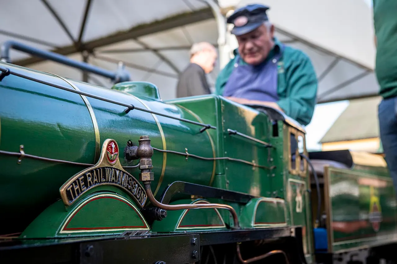 A man in a cap and coloured jacket stands on a vintage green steam locomotive marked "The Railwayman's Shed" under a covered platform.