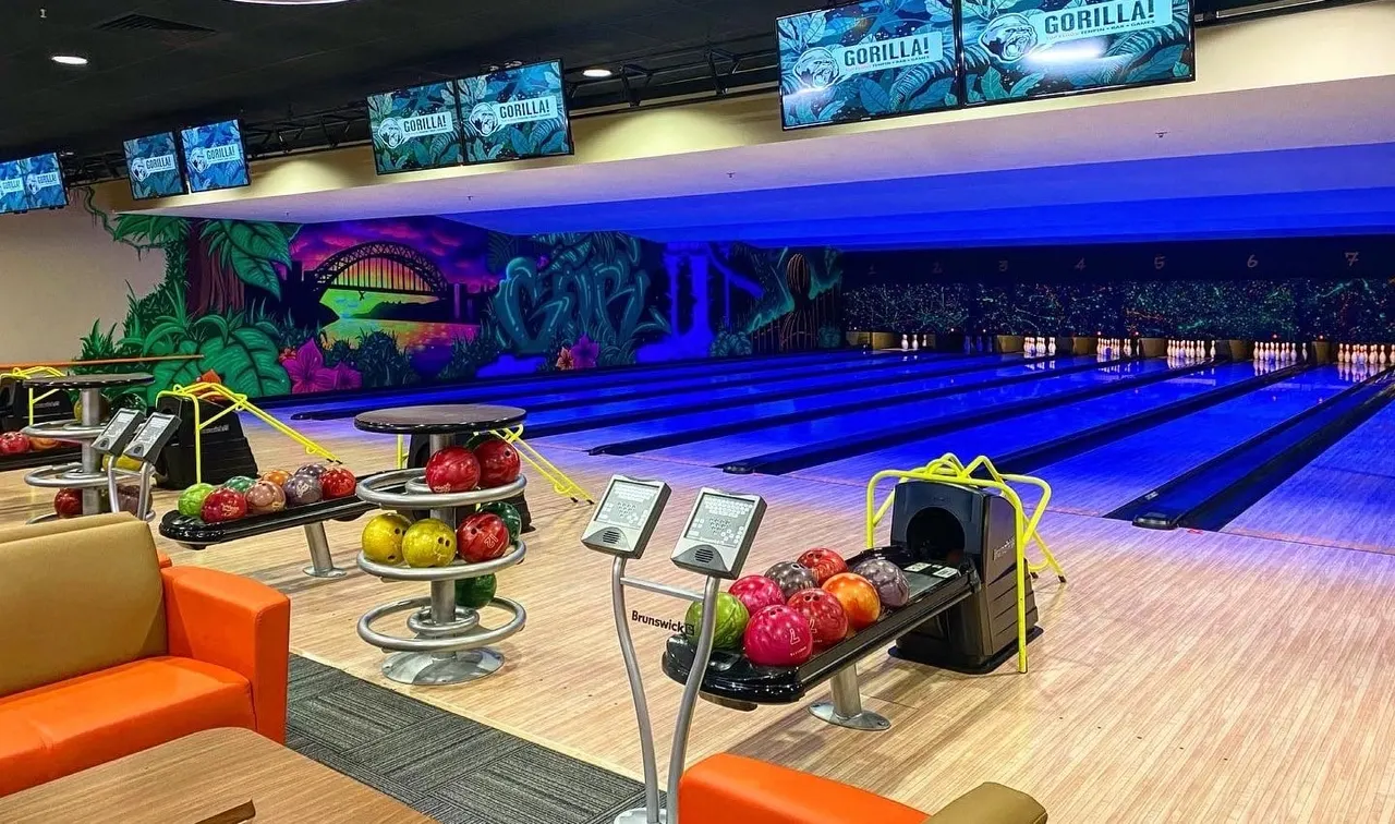 Modern bowling alley interior with glowing blue lanes, colorful bowling balls on stands, and vibrant jungle-themed wall murals under blue lighting.