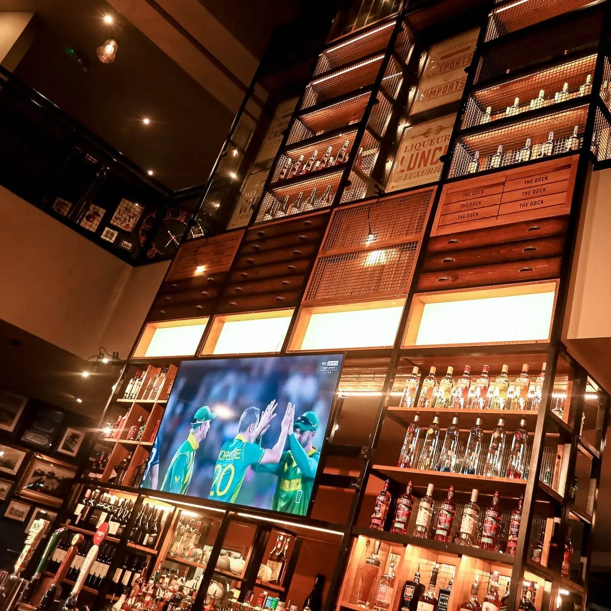 Interior of a sports bar with wooden shelving displaying bottles of spirits and liquor, a TV screen showing cricket players, and warm ambient lighting.