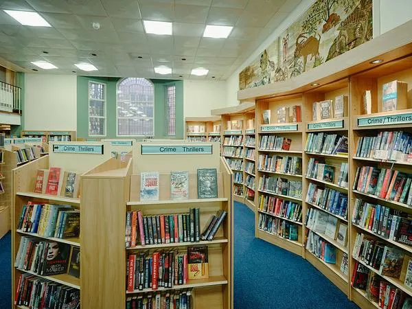 Library interior with wooden bookshelves displaying books, blue carpet flooring, and decorated ceiling panels.