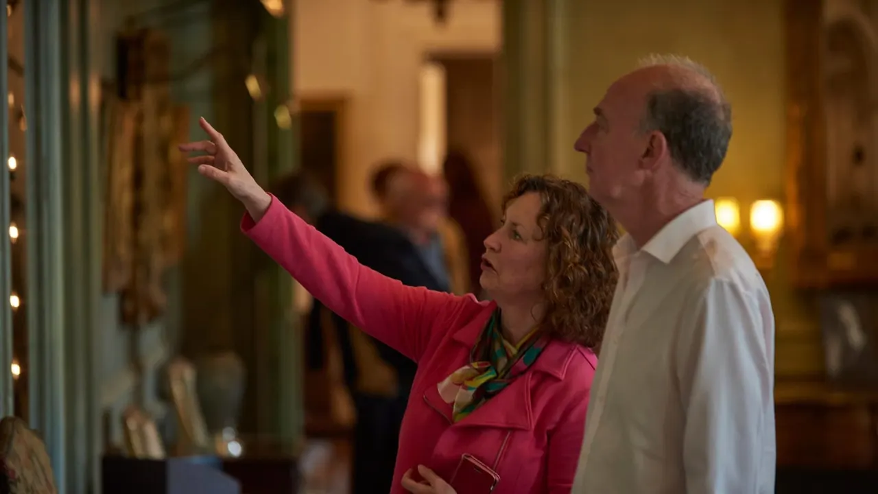 Woman in pink jacket gives guided tour to man in white shirt inside illuminated historic interior.