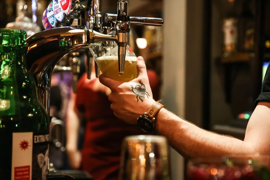 Bartender pouring a draught beer from a tap into a glass at a busy pub.