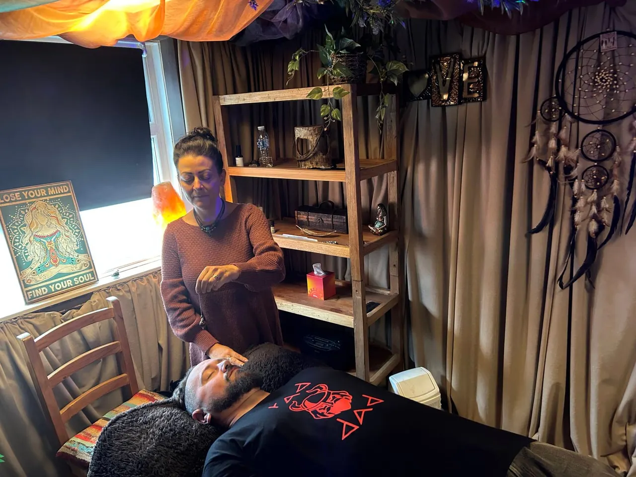 A tarot reader performs a reading for a client lying on a table in a dimly lit room decorated with mystical objects and dreamcatchers.