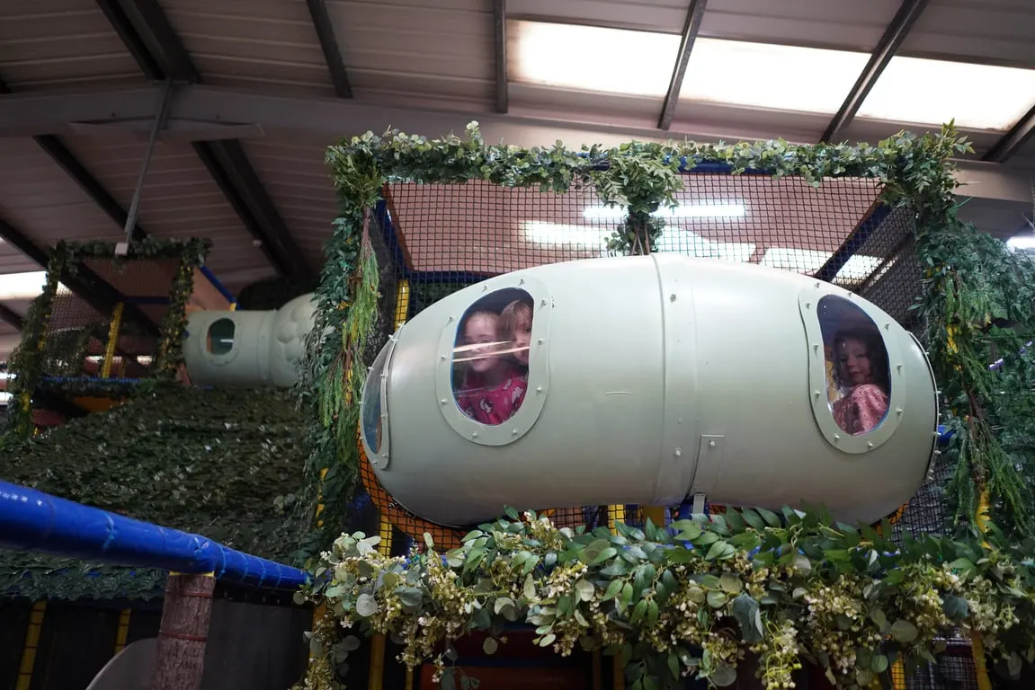 Two children looking out of circular windows in a white capsule-shaped play structure decorated with artificial foliage indoors.