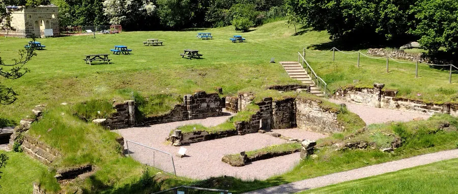 Elevated view of Roman fort ruins with picnic tables scattered across grassy grounds and wooded surroundings.