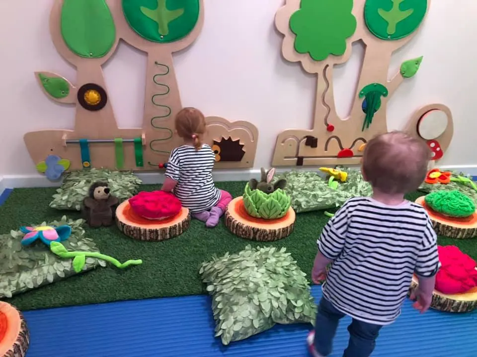 Two toddlers in striped shirts exploring a colourful interactive garden play installation with fabric plants and wooden tree structures.