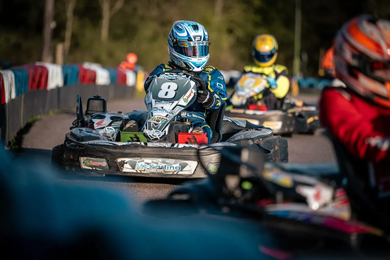 Go-kart racer in blue helmet and number 8 kart on outdoor track with other competitors and coloured barriers.