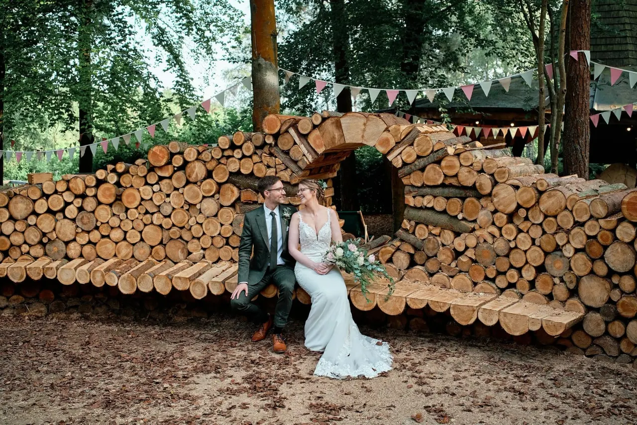 Bride and groom sit together on stacked logs at a woodland venue, with bunting and trees in the background.