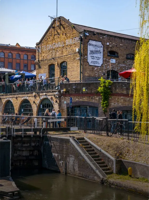 Historic brick building with outdoor terraces beside a canal, visitors seated under red umbrellas on a sunny day.