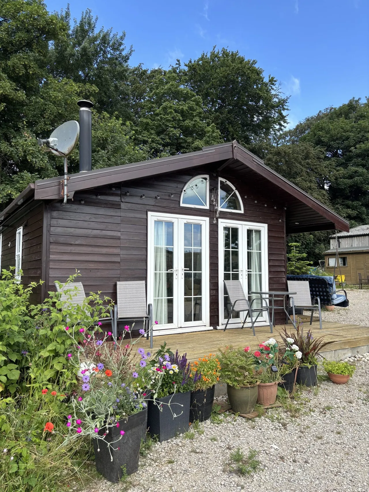 Modern dark timber lodge with decking, white doors and windows, surrounded by flowering plants and garden containers.